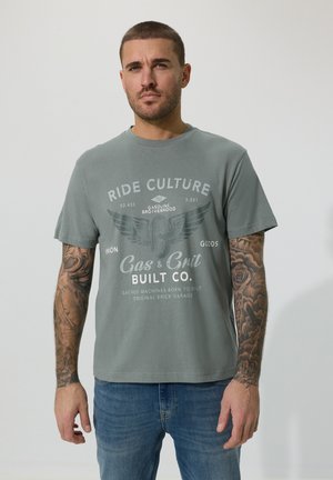 Homme aux cheveux courts et tatouages sur les deux bras, portant un t-shirt graphique gris et un jean bleu, debout devant un fond clair uni.