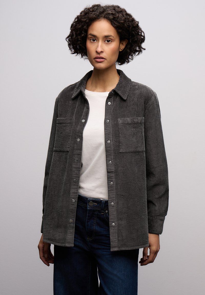 Grijs corduroy overhemd met knopen en twee borstzakken, gecombineerd met een wit T-shirt en donkere denim jeans. Zachte textuur met duidelijke verticale lijnen.