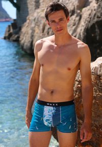 Maillots de bain bleus avec un motif aquatique, dotés d'une ceinture élastique noire portant l'inscription "WAXX". Le tissu semble lisse et bien ajusté.