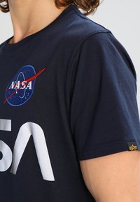 Camiseta azul marina de algodón con cuello redondo, que presenta el logo blanco de "NASA" y el texto "5A". Incluye una pequeña etiqueta de tela con acento.