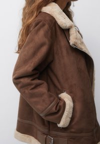 Brun faux suede jakke med en beige faux pelskrave. Funktioner inkluderer sidelommer og spænde detaljer ved taljen. Blød tekstur hele vejen igennem.