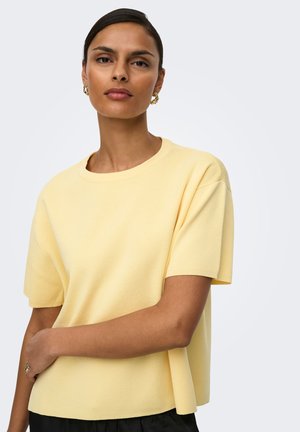 Donna che indossa un top lavorato a maglia giallo chiaro a maniche corte con scollo rotondo, orecchini a cerchio in oro e un anello d'oro sulla mano sinistra.