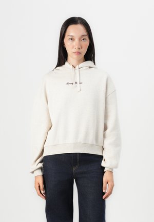 Light beige cropped hoodie with a drawstring hood, featuring embroidered text in dark brown. Paired with dark jeans. Smooth texture.