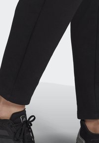 Benen in zwarte cropped broeken boven zwarte Adidas sneakers met veters, staande op een lichtgrijze vloer.