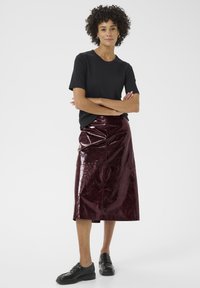 Une femme aux cheveux bouclés se tient avec confiance, portant un t-shirt noir et une jupe bordeaux brillante, assortie de chaussures noires, les bras croisés.