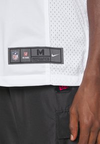 Primer plano de una camiseta de malla blanca de la NFL con etiqueta de tamaño M y logotipos, usada por una persona de piel oscura. También se muestran pantalones negros con un logotipo de gorra roja.