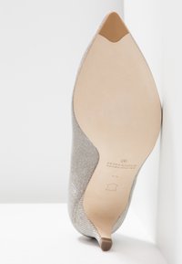 Chaussure pointue en textile argenté avec une finition texturée, un talon fin et une semelle beige, avec le nom de la marque embossé au dos.