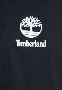 Melna kokvilnas T-kreklu ar baltu Timberland logo un koku simbolu, ar gludu tekstūru un klasisku apaļu kakla dizainu.
