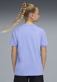 Νεαρός με μακριά πλεγμένα αλογοουρά, ντυμένος με ανοιχτό μοβ κοντομάνικη μπλούζα και μαύρο παντελόνι, στέκεται με την πλάτη γυρισμένη προς το γκρι φόντο.