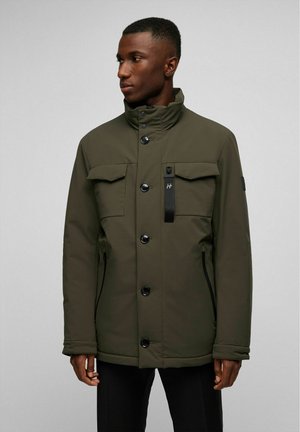 Olivgrüne isolierte Jacke mit hohem Kragen, großen Fronttaschen, Knopfschluss und seitlichen Reißverschlusstaschen. Strukturiertes Gewebe.