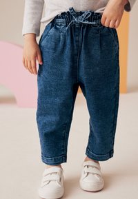 Pantaloni di denim con cintura elastica e coulisse, caratterizzati da una vestibilità rilassata, tasche laterali e un lavaggio blu scuro. Scarpe bianche semplici.