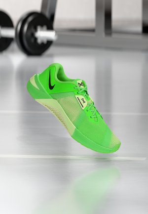 Jasnozielony sportowy sneaker z czarnym logo Nike zawieszony nad podłogą siłowni w pobliżu sprzętu do podnoszenia ciężarów.