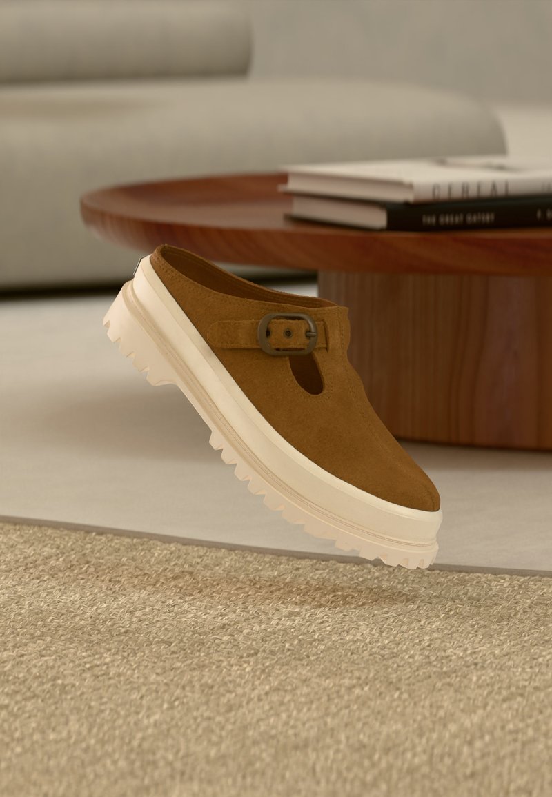 Καφέ σουεντ παπούτσι τύπου slip-on με λευκή παχιά σόλα και πλαϊνή αγκράφα, τοποθετημένο κοντά σε στρογγυλό ξύλινο τραπεζάκι καφέ με στοίβες βιβλίων στο φόντο.