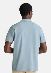 Muž s krátkými kudrnatými vlasy, oblečený v světle modrém polo tričku s krátkým rukávem, stojící zády k fotoaparátu na bílém pozadí.