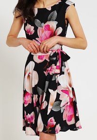 Ärmlös svart klänning med stora rosa och vita blommönster, figursydd midja knuten med ett matchande bälte i tyg.