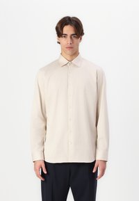 Chemise beige clair à manches longues avec un col, fermeture à bouton, et texture de tissu douce. Associée à un pantalon sombre, la tenue est minimaliste.