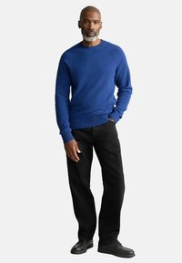 Blauer Rundhalsausschnitt-Pullover aus weichem Strick, kombiniert mit schwarzen geraden Jeans und schwarzen Schuhen. Model steht mit den Händen in den Taschen.