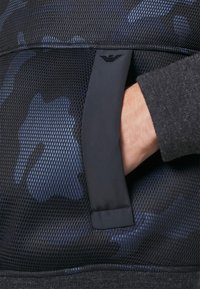Main dans la poche latérale d'un vêtement texturé noir et bleu avec un petit logo noir au-dessus de l'ouverture de la poche.