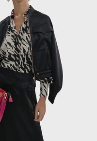 Black bomber jacket with a zip front, featuring a patch pocket. Below, a zebra-print blouse with pleats, paired with a black skirt and pink handbag.