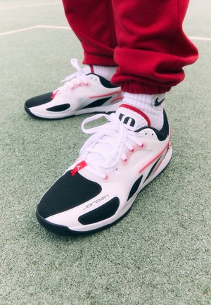 Baltas un melns sporta apavos ar sarkanām akcentiem, ar sietveida augšdaļu, perforācijām un redzamām zīmola atzīmēm sānos. Apavi ir saistīti.