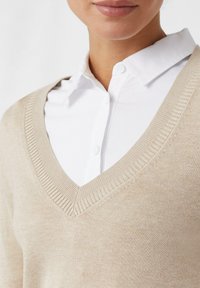 Frau in einem beigen V-Ausschnitt-Pullover über einem weißen Hemd, die Schultern, den Hals und den unteren Teil des Gesichts vor einem weißen Hintergrund zeigt.