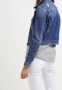 Žena v modré džínové bundě přes šedé tričko a bílé kalhoty, zobrazená zezadu a z boku na jednobarevném pozadí.