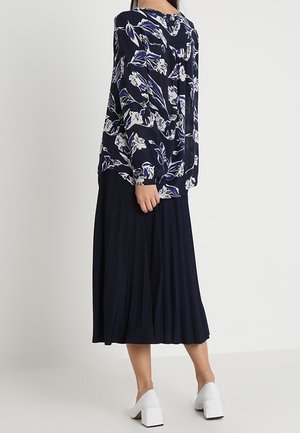 Femme portant un chemisier à manches longues à motif floral foncé, une jupe midi plissée bleu marine et des chaussures blanches à talons blocs, debout de profil sur un fond uni.