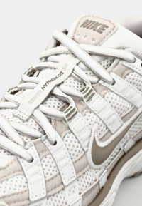 Nike-Laufschuhe mit einem Obermaterial aus weißem und hellem beigem Mesh, metallischen Akzenten und einer strukturierten Sohle, sowie Schnürsenkeln und Brandingdetails.
