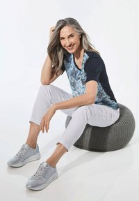 Lichtblauw en marineblauw bloemenblouse, witte broek en grijze sneakers, zittend op een textuur grijze poef tegen een witte achtergrond.