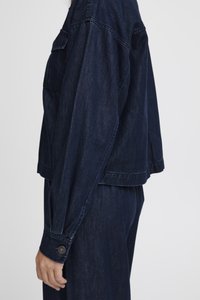 Σκούρο μπλε τζιν μπουφάν με χαλαρή εφαρμογή, ορατές ραφές, ρυθμιζόμενους μανσέτες με κουμπιά και τσέπη στην αριστερή πλευρά.