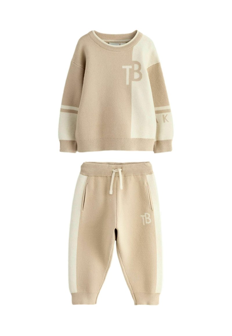 Beige gebreide pullover met witte accenten en "TB" letters; bijpassende beige joggingbroek met witte zijpanelen en een trekkoord in de taille.