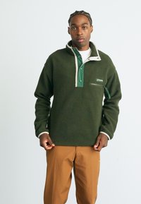 Man with braided hair wearing green fleece pullover with buttoned collar and tan pants, standing against plain light background.