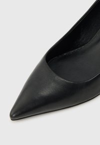 Chaussure plate en cuir noir à bout pointu avec une texture lisse et un empeigne basse, présentée sur un fond clair uni.