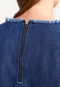 Vêtement en denim présentant un col effiloché, des coutures visibles et une fermeture éclair dorée à l'arrière. Tissu bleu avec une texture lisse.