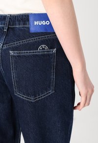 Tamno plave traperice s plavom kožnom oznakom na kojoj piše "HUGO." Imaju stražnji džep s kružnim "HGO" logom i kontrastnim bijelim šavovima.