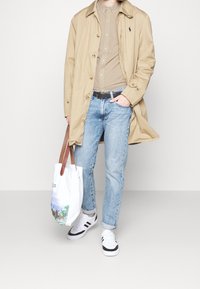 Beige trenchcoat, ruudullinen paita, vaaleansiniset farkut, valkoiset lenkkarit mustilla yksityiskohdilla ja valkoinen kangaskassi, jossa on maisemakuva.