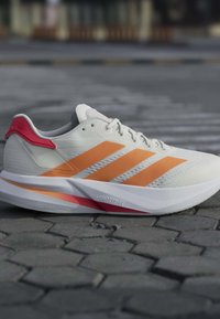 Chaussure de course blanche avec des bandes orange et un détail rouge au talon sur un sol pavé en pierre grise, soulignant le design amorti de la semelle Lightstrike.