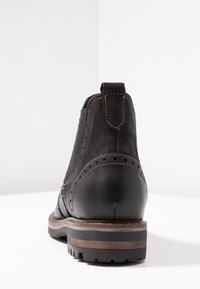 Černá kotníková bota s kombinací semišového a koženého svršku, zdobená brogue vzorem, hnědým šitím a pevnou gumovou podrážkou s dřevěným akcentem.