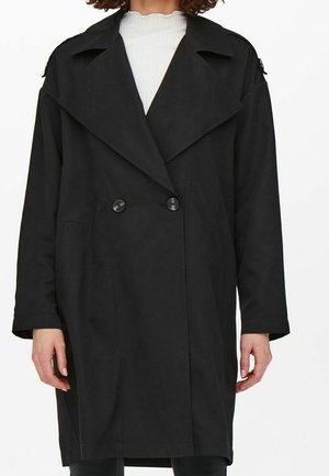 Person iført en lang, sort dobbeltradet frakke med brede revers over en hvid top med høj hals, hænderne afslappet ved siderne.