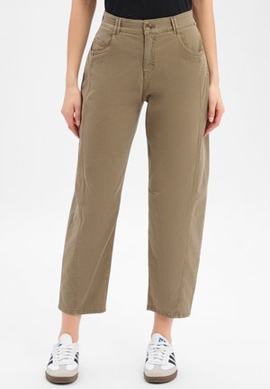 Woman wearing cropped khaki pants and white sneakers with black stripes, standing against plain light background.