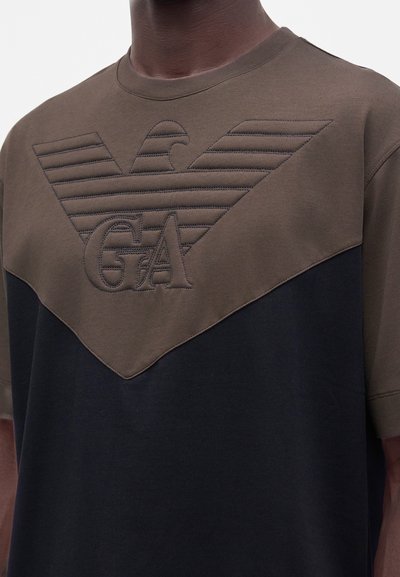 Άνδρας που φοράει δίχρωμο καφέ και μαύρο T-shirt με μεγάλο ανάγλυφο λογότυπο αετού και τα γράμματα GA στο στήθος.