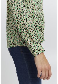Camicia a maniche lunghe realizzata in un tessuto beige chiaro, caratterizzata da un motivo astratto verde, nero e bianco. I bottoni sui polsini sono di un verde chiaro.