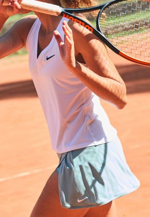 Un débardeur de sport blanc avec une texture côtelée et un décolleté plongeant, associé à un short de tennis bleu clair avec un logo sur le côté. Le fond est un court en argile.