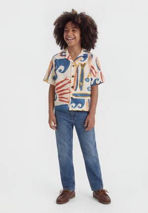 Niño sonriente con cabello rizado que lleva una camiseta de manga corta con formas abstractas en azul y naranja, pantalones vaqueros azules y zapatos marrones.