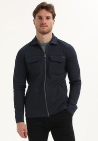Chaqueta con cremallera en color navy, de tela suave, con dos bolsillos en el pecho y cuello, que presenta una textura lisa y un diseño ajustado.