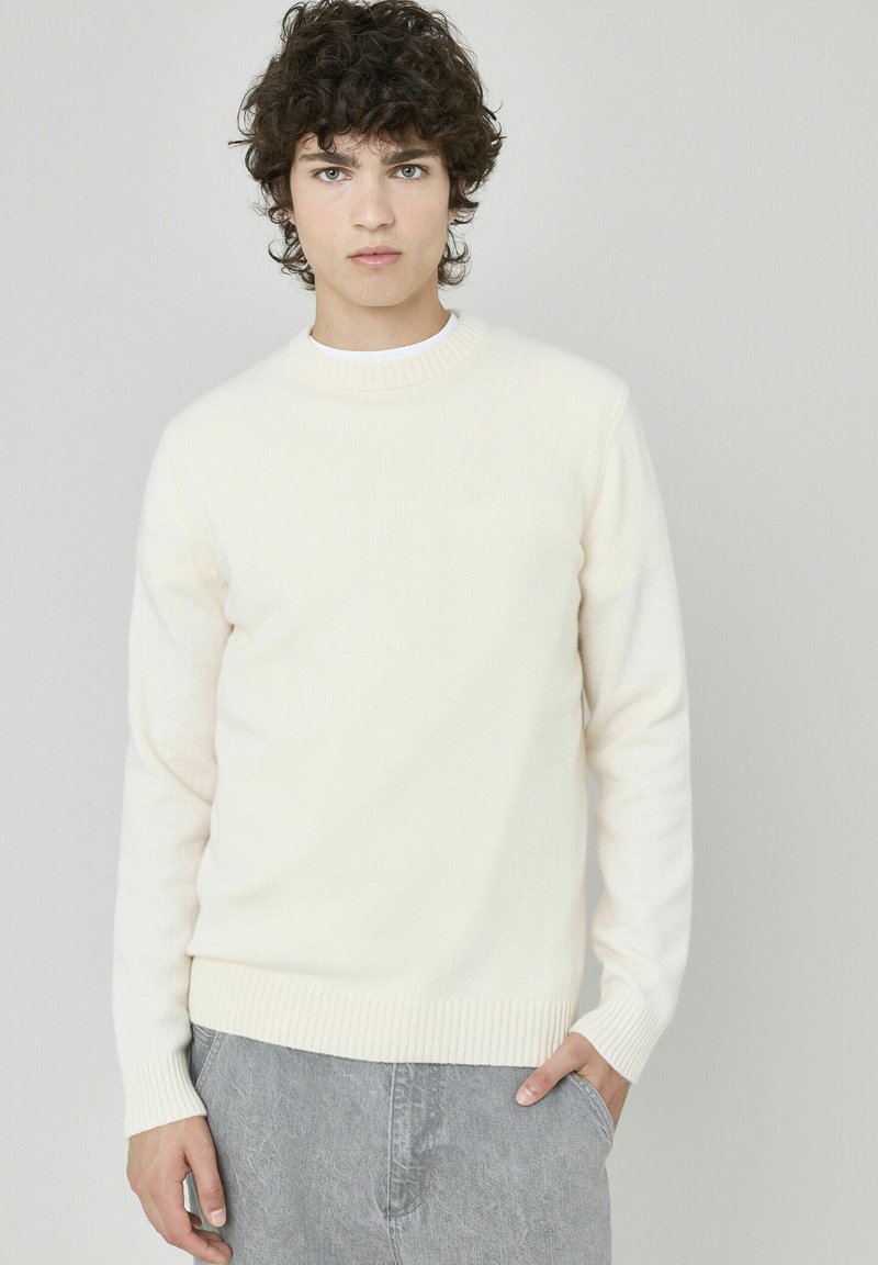 Pull en maille couleur crème avec un col rond, des poignets et un ourlet côtelés ; présente une surface texturée subtile. Porté sur un pantalon gris.