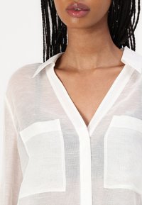Blusa bianca semi-trasparente con colletto, pattina dei bottoni e due tasche frontali. Tessuto testurizzato con strisce sottili e finitura liscia.