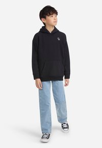 Junge trägt einen schwarzen Kapuzenpullover, hellblaue Jeans und schwarze Turnschuhe, steht da und schaut zur Seite vor einem weißen Hintergrund.