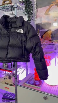 Geacă puffer neagră cu un design cropped, având închidere cu fermoar, secțiuni mari matlasate și un logo alb pe partea stângă a pieptului.