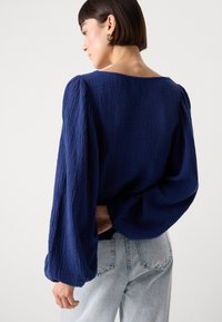 Navy blue textured blouse with long, puffed sleeves and a wide neckline, paired with light blue high-waisted jeans.